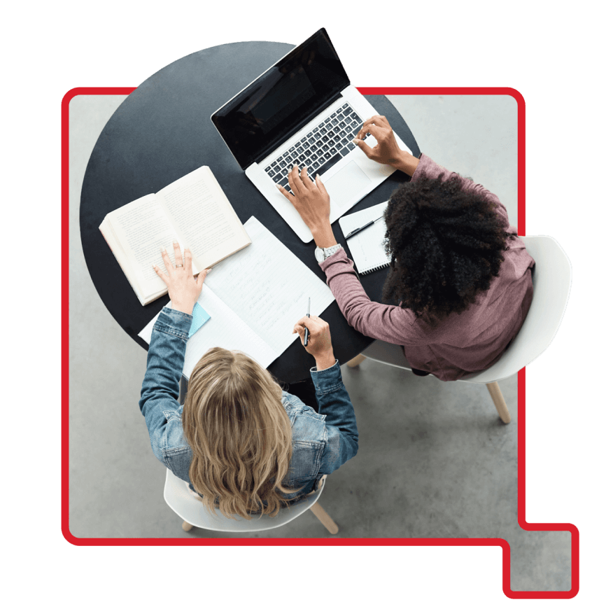 Two women working together at a round table with a laptop, notebook, and open book, viewed from above.