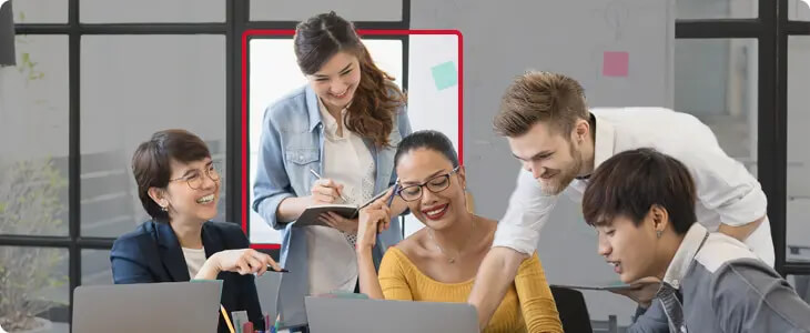 Five colleagues of diverse backgrounds collaborating and smiling around laptops in a modern office setting.